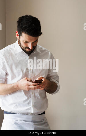 Half length portrait of a bearded man wearing a white apron, using his smart phone. Stock Photo