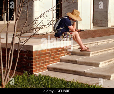 A young man in casual attire sits on the stairs outside a building reading text messages or emails on a cell phone. Stock Photo
