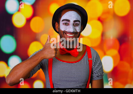 Pantomime man with facial paint posing for camera interacting giving thumbs up smiling, blurry lights background Stock Photo