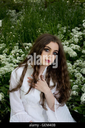 Beautiful woman wearing a long white dress sitting in white flowers looking straight to camera Stock Photo