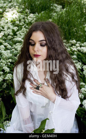 Beautiful woman wearing a long white dress sitting in white flowers with her eyes closed Stock Photo