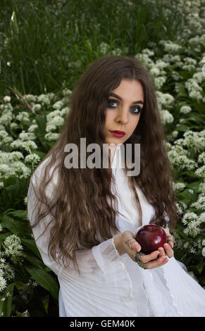 Beautiful woman wearing a long white dress sitting in white flowers holding a red apple Stock Photo