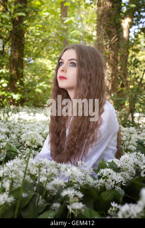 Beautiful woman wearing a long white dress sitting in white flowers looking into the distance Stock Photo