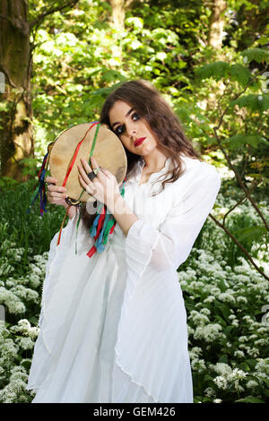 Beautiful woman wearing a long white dress standing in a forest holding a tambourine Stock Photo
