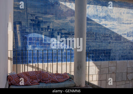 A homeless man sleeps under blankets in front of Azulejo tiles in a city park, Lisbon Portugal. Stock Photo