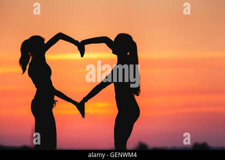 Landscape with silhouette of young sporty women holding hands in heart shape on the background of colorful sky at sunset in summ Stock Photo