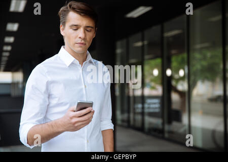 Serious young businessman standing and using smartphone near business center Stock Photo
