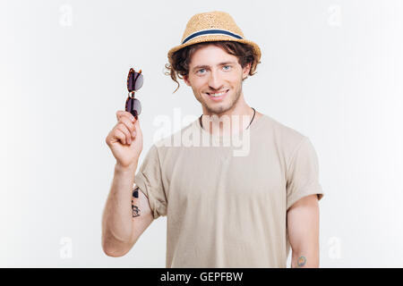 Cheerful confident young man in hat holding sunglasses over white background Stock Photo