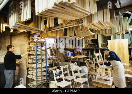 Two men standing in a carpentry workshop, one working on a wooden armchair. Stock Photo