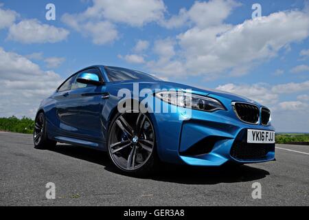 low down shot of a BMW M2 standing on a road with trees in the middle distance and clouds against a blue sky above Stock Photo