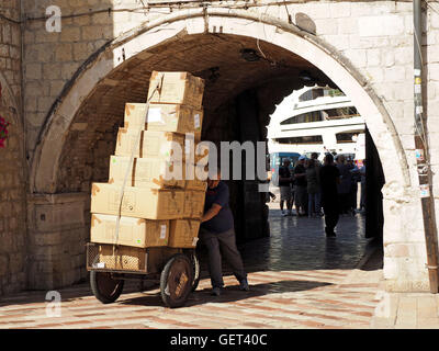 delivery worker pushing barrow piled high with cardboard boxes through archway into marbled square in Old Town Kotor, Montenegro Stock Photo