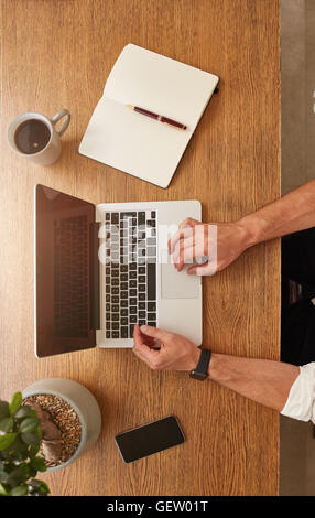 Top view of man using a laptop on a wooden table, a coffee cup, diary, mobile phone and potted plant on work desk. Stock Photo