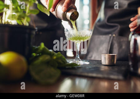 Close up shot of bartender hand pouring basil cocktail in a glass from shaker. Barman preparing classic modern basil smash cockt Stock Photo