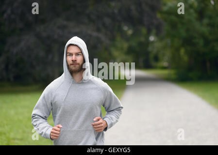 Young man wearing a hoodie jogging through a park in a health and fitness concept with copy space Stock Photo