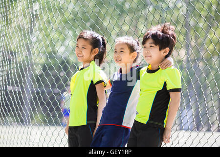 Happy Chinese children in sportswear leaning against chainlink fence Stock Photo