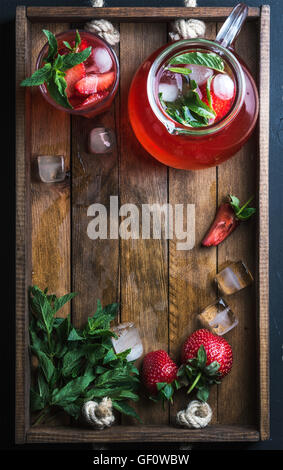 Homemade strawberry lemonade, mint and ice, served with fresh berries on rustic wooden tray over dark background, top view, copy space Stock Photo