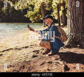 Portrait of senior man sitting near a lake using compass for searching direction. Mature man hiking in nature using compass for Stock Photo
