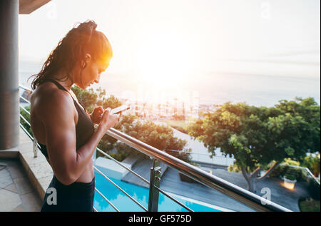 Side view of fitness woman standing in balcony and using mobile phone. Healthy female reading text message on the mobile phone. Stock Photo