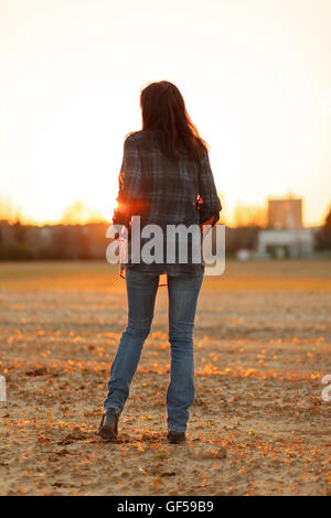Young woman with long hair from behind walking on a plowed land in the sunset Stock Photo