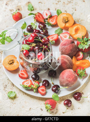 Healthy summer fruit variety. Sweet cherries, strawberries, peaches, apricots and mint leaves on white ceramic serving plate over light concrete background. Top view, selective focus Stock Photo