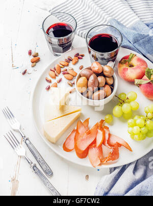 Wine snack set. Cheese, chicken carpaccio, mediterranean olives, fruits, nuts and two glasses of red on ceramic plate over white wooden background Stock Photo