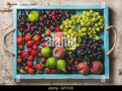 Healthy summer fruit variety. Black and green grapes, strawberries, figs, sweet cherries, peaches in blue wooden tray over light concrete background Stock Photo