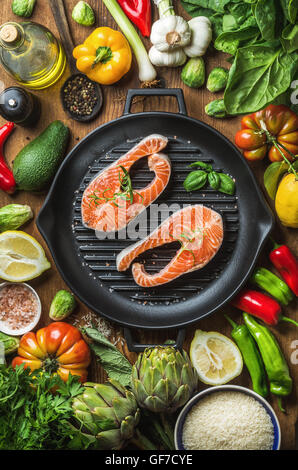 Dinner cooking ingredients. Raw uncooked salmon with vegetables, rice, herbs, lemon, artichokes, spices in iron grilling pan over wooden background, top view Stock Photo