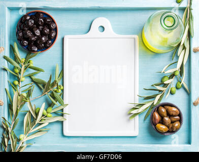 Two bowls with pickled green and black olives, olive tree sprigs, oil, white ceramic board in center. Copy space. Stock Photo