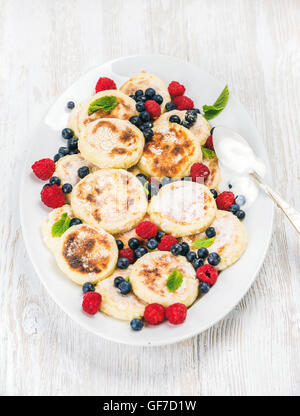 Syrniki or cottage cheese pancakes with fresh forest berries and sour cream sauce in serving dish over white wooden background Stock Photo