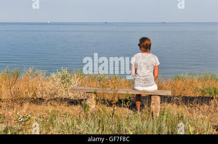 white middle aged woman with short hairs in t-shirt, shorts and sunglasses sitting on old wooden bench with Black Sea seascape Stock Photo