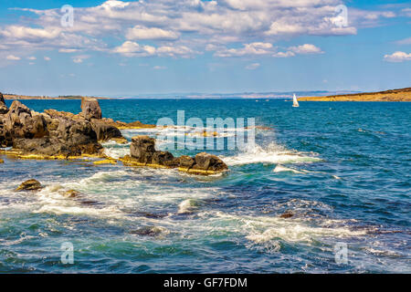Small boat sails at distance in the sea. waves break on the huge boulders  on the rocky shore. on the other side of of seascape Stock Photo