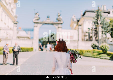 Gorgeous stylish red headed bride in vintage white dress walking in old city, view from behind Stock Photo