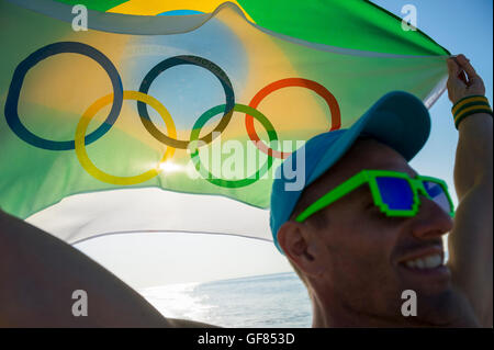 RIO DE JANEIRO - MARCH 10, 2016: Smiling athlete holds Olympic and Brazil flags over the beach shore at sunrise. Stock Photo