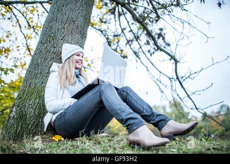 MODEL RELEASED. Young woman sitting at tree trunk and using laptop. Stock Photo
