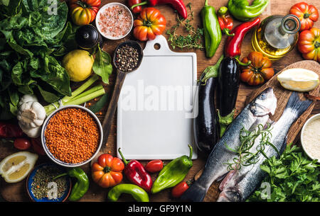 Ingredients for cooking healthy dinner. Raw uncooked seabass fish with vegetables, grains, herbs and spices over rustic wooden background, white ceramic board in center Stock Photo