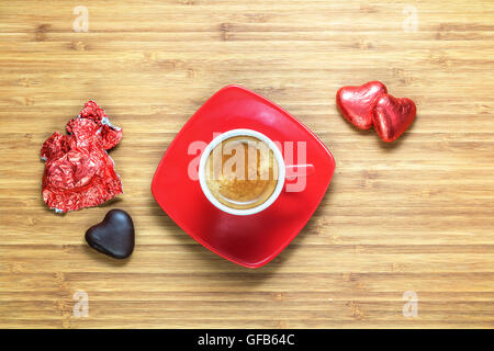 Heart shaped sweets wrapped in a bright red foil lying on wooden texture with  cup of coffe near it. Background for romantic themes. Stock Photo