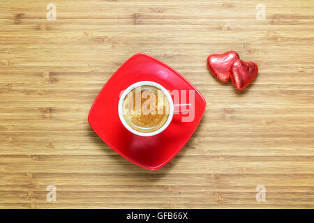 Heart shaped sweets wrapped in a bright red foil lying on wooden texture with  cup of coffe near it. Background for romantic themes. Stock Photo