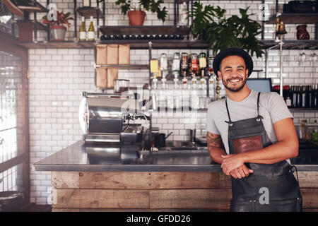 Portrait of young man standing at the counter in his cafe. Coffee shop working in apron and hat smiling at camera. Stock Photo