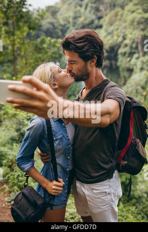 Loving young couple kissing and taking self portrait with their cell phone. Couple in love taking self picture in forest. Stock Photo