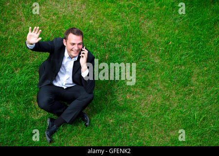 Cheerful young businessman calling by phone sitting on green grass Stock Photo