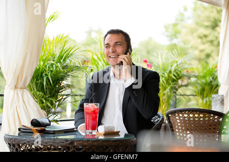 Young businessman talking on a cell phone and drinking a cocktail sitting in a summer restaurant Stock Photo