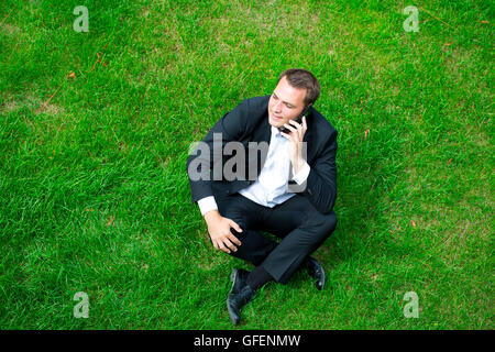 Cheerful young businessman calling by phone sitting on green grass Stock Photo