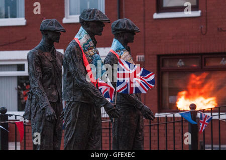 Belfast, Northern Ireland. 11 Jul 2016 - The reflection of a bonfire in a window behind a public art display remembering the shipyard workers of Belfast, which has been decorated with Orange 'Sashes' or collarettes and Union Flags in readiness for the 12th July celebrations