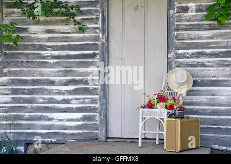 Vintage camera on old suitcase by wooden door with hat and flowers on wicker chair. Stock Photo