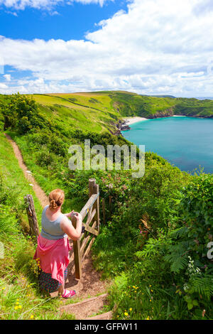 A woman overlooking Lantic Bay on a summers day in hot sunny weather as she opens a gate to continue her walk along the Coastal Path Stock Photo