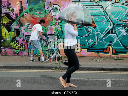London, UK. 1st August 2016 A girl with a see trouhg umbrella walked pass in front of some street artist who ignored the rain and got on with their work. @ Paul Quezada-Neiman/Alamy Live News Stock Photo