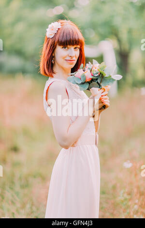 Young red-haired bride in a wreath is standing holding bouquet of pink flowers Stock Photo