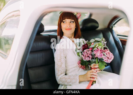 Portrait of beautiful red-head bride with pink bouquet in hands sitting in the car and looking aside Stock Photo