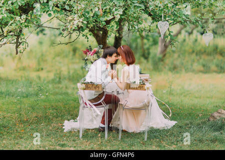 Bride, groom sitting in forest, view from behind. The two transparent chairs decorated with signs for her and him Stock Photo