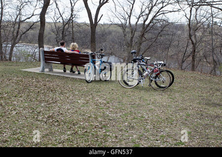 Bicycling couple resting on park bench overlooking the Mississippi River. Mississippi River Boulevard, St Paul Minnesota MN USA Stock Photo
